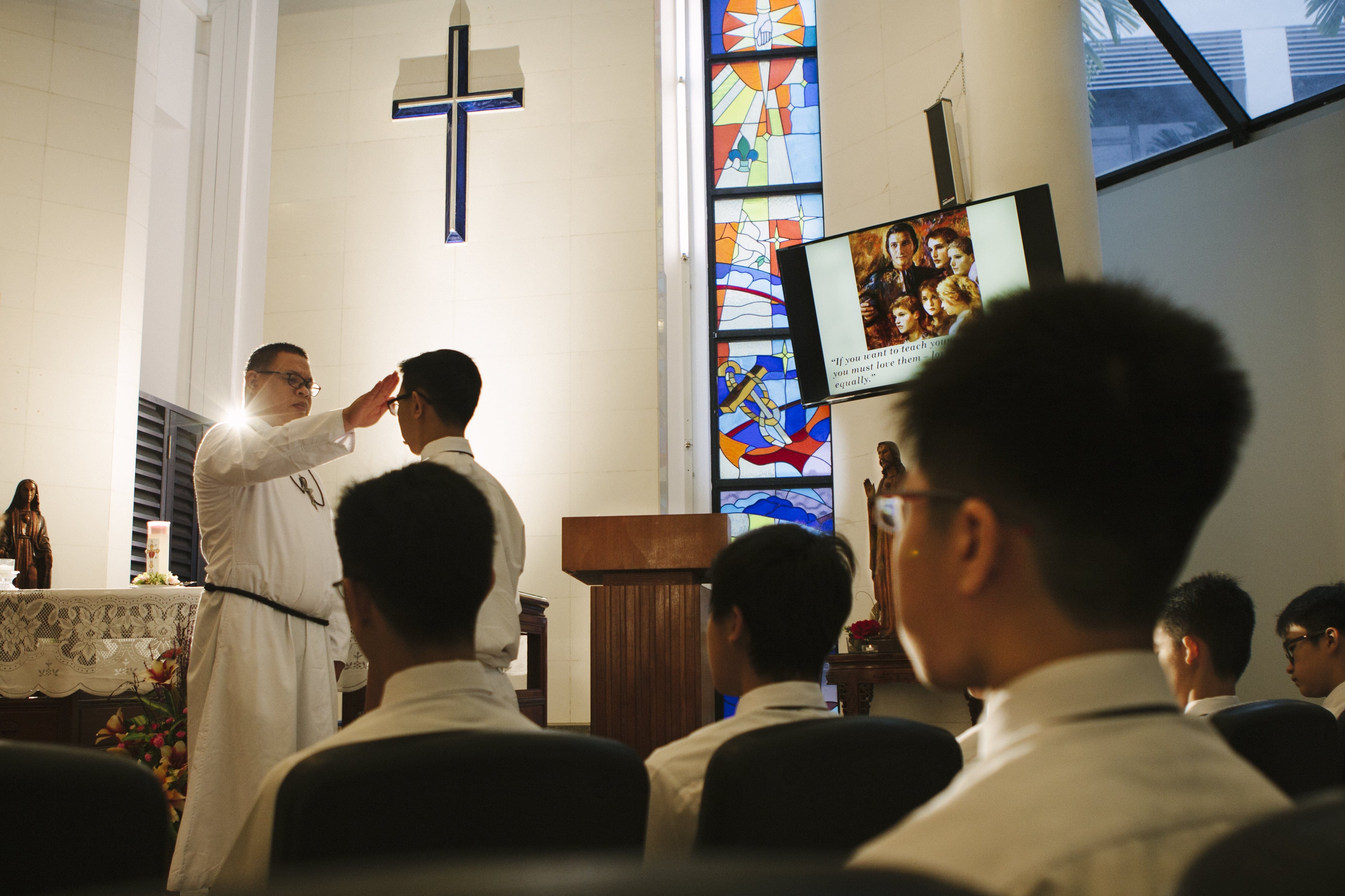 A priest in white robes blesses a boy before seated students in a chapel with a cross and stained glass window.